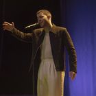 Ein Mann singt leidenschaftlich in ein Mikrofon, trägt eine dunkle Jacke und helle Hose, der Hintergrund ist blau beleuchtet.