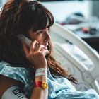 Frau im Krankenhausbett, telefoniert, trägt Krankenhausarmbänder und ein gepunktetes Kleid, wirkt konzentriert.