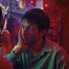 "Aperol"-Flasche links, junger Mann in grünem Hemd, stützt Kopf, bunte Partystimmung.