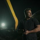 Ein Mann in dunkler Kleidung steht nachts neben einem Autowrack, hält ein Objekt, beleuchtet von hellem Scheinwerferlicht.