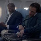 Keine sichtbaren Texte. Zwei Männer sitzen nebeneinander in einem Flugzeug, einer benutzt ein Handy, der andere durchsucht eine Tasche.