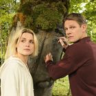 OTTO, Polly, Mia sind in einen Baum geritzt. Zwei Personen stehen dort; eine blickt nachdenklich, die andere schnitzt.