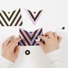 Hände arrangieren geometrische Formen aus Papier mit schwarzen und schillernd violetten Streifen auf weißem Untergrund.