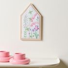 Illustration von bunten Blumen in einem hausförmigen Holzrahmen, daneben zwei rosa Kerzenhalter auf einem Tisch.