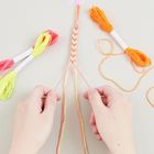 Hände flechten bunte Fäden zu einem Armband. Vier Garnfarben: neonpink, gelb, orange, beige.