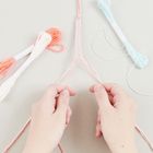 Zwei Hände flechten rosa und weiße Bänder. Daneben liegen Garnbündel in Weiß, Rosa und Hellblau.