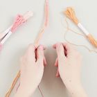 Zwei Hände flechten rosa und orange Fäden, umgeben von Garnsträngen.