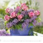 "Edelrosen 'Varience', Flieder und Herz aus Efeu." Ein Blumenarrangement in Rosa und Lila auf einem Tisch.