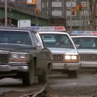 Zwei Polizeiautos mit blinkenden Lichtern verfolgen ein schwarzes Auto auf einer rauen Straße in städtischer Umgebung.