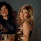 Zwei lachende Frauen in Partykleidung; eine mit dunklem, die andere mit blondem Haar; rechts eine Lampe.