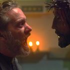 Ein Mann in Priesterkleidung schreit ein Kruzifix an, das von Kerzenlicht beleuchtet wird.