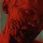 Keine Texte. Eine rote, verstörende Figur mit verzerrtem Gesicht, im Hintergrund verschwommene grüne Lichter.