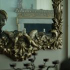 Ein Puppengesicht mit gruseligen, leer wirkenden Augen spiegelt sich in einem prunkvollen goldenen Spiegelrahmen.