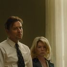 Drei Personen in förmlicher Kleidung vor einem Fenster, ernste Gesichtsausdrücke. Langer, heller Vorhang im Hintergrund.