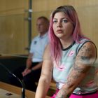 Eine Frau mit rosa Haaren sitzt vor Glas. Sie trägt ein Oberteil mit rosa Lippen und hat tätowierte Arme. Im Hintergrund ein sitzender Mann.