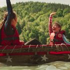Kinder in roten Schwimmwesten auf einem Floß, Hände erhoben, vor grünem Wald und blauem Himmel.