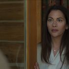 Eine Frau schaut ernst aus einem Holzrahmen-Fenster in einem gemütlichen Raum mit Holzverkleidung.