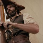 Zwei Männer auf einem Segelschiff. Der bärtige Mann trägt einen Dreispitz, und ein Mast mit Segeln ist sichtbar.
