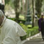 Ein älterer Mann in weißer Kleidung und mit goldener Halskette blickt nach hinten. Im Hintergrund gehen Personen in religiöser Kleidung.
