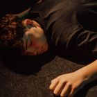 Ein Junge liegt auf dem Boden, sein Gesicht seitlich, mit geschlossenen Augen. Er trägt ein dunkles Shirt, Beleuchtung ist schummrig.