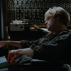 Neben einem leuchtenden Bildschirm mit animierten Figuren sitzt eine Person in einem Stuhl, umgeben von Musik-Equipment.