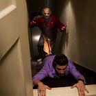 Ein Mann kriecht in Angst eine Treppe hinauf, während hinter ihm eine maskierte Person mit einem Haken steht.