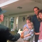 Ein Mann mit Hut gestikuliert zu einer besorgten Familie auf einer Veranda: Frau, Mann, Junge und Mädchen mit Teddybär.