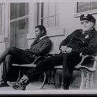 Zwei Männer sitzen auf einer Bank. Der eine trägt einen Anzug, der andere eine Polizeiuniform mit Sonnenbrille.