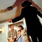 Ein Mann im schwarzen Anzug und Hut wirft einen großen Schatten auf eine verängstigte Gruppe von vier Personen an einer Tür.