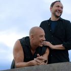 Zwei Männer lächeln und stehen nah beieinander draußen unter blauem Himmel. Einer stützt sich auf eine Mauer.