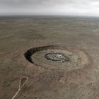 Eine weite, karge Landschaft mit einem großen, kreisförmigen Krater in der Mitte, umgeben von flachem, bewachsenem Terrain.