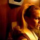 Eine Frau mit blondem Haar und Haarreif in einem holzgetäfelten Raum. Warmes Licht, ernster Blick nach rechts.