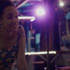 Eine Frau mit Blumen-Tattoo, hellem Blumenkleid, steht in bunter, beleuchteter Bar und lächelt jemanden an.