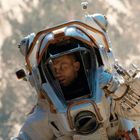 Ein Astronaut in einem Raumanzug, er blickt aus dem Helm, vor einem verschwommenen, erdigen Hintergrund.
