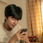 Ein junger Mann lächelt auf sein Handy. Im Hintergrund sind Vorhänge, ein Ventilator und Topfpflanzen zu sehen.