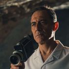 Ein Mann in weißem Hemd hält eine alte Filmkamera und schaut ernst, mit dunklem Hintergrund.