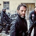 Drei Männer in Lederjacken, stehen vor alten Motorrädern. Einer trägt einen Helm und die Umgebung wirkt rustikal.