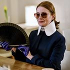 Eine stilvolle Frau mit Sonnenbrille und großem, weißen Kragen hält einen schwarzen Fächer und trägt lila Handschuhe.