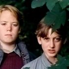 Zwei junge Menschen in Outdoor-Kleidung stehen vor dichten grünen Blättern. Sie schauen ernst in die Kamera.