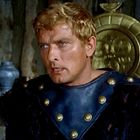 Ein Mann in antiker Rüstung mit blonden Haaren, ernstem Blick; im Hintergrund ein dekorativer Schild und eine Vase.