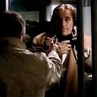 Ein Mann in einem hellen Mantel steht einer anderen Person gegenüber und greift zu deren Mantel in einer Telefonzelle.