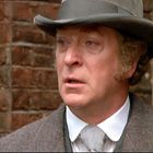 Ein Mann mit ernster Miene trägt einen grauen Hut und Mantel, vor einer Ziegelmauer stehend.