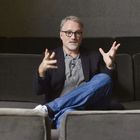 Keine sichtbaren Texte. Ein Mann mit Brille sitzt auf einem Sofa, trägt ein Sakko und Jeans und gestikuliert mit den Händen.