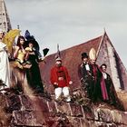 Ich kann den Text nicht erkennen. Mehrere Menschen in Kostümen und mit Hüten stehen auf einer Mauer vor einem Gebäude.