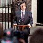 Ein Mann in Anzug steht ernsthaft hinter einem Redepult mit Mikrofon, draußen vor einem Gebäude mit schwarzer Tür.