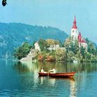 Zwei Personen in einem Boot auf einem See vor einer bewaldeten Insel mit einer Kirche und roten Dächern.