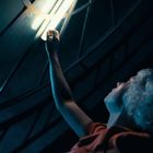 Keine Texte sichtbar. Eine Person mit lockigem Haar streckt ihre Hand zu einem leuchtenden Fenster hinauf.