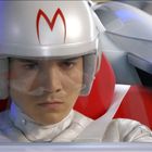 Ein silberner Helm trägt ein rotes "M". Junge Person sitzt konzentriert in einem futuristischen Cockpit auf einem roten Sitz.