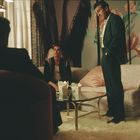 Männer in Anzügen in einem Wohnzimmer mit floralem Hintergrund, einem Glastisch und Essen in chinesischen Imbiss-Boxen.