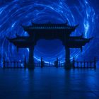 Ein traditionelles asiatisches Tor steht vor einem wirbelnden blauen dimensionalen Portal, flankiert von zwei Personen.